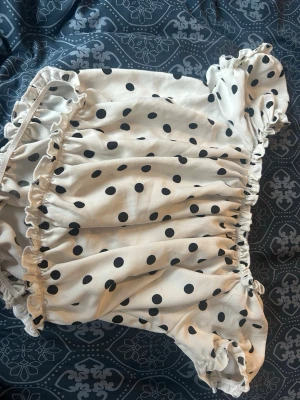 Prickig puffärm blus med volang - Supersöt blus i beige med svarta prickar och puffiga korta ärmar. Toppen har volangdetaljer längs kanterna och en söt, romantisk känsla. Perfekt för dig som gillar trendiga och lekfulla plagg.