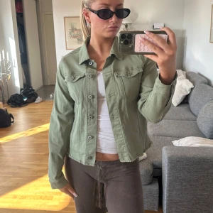 Grön jeansjacka med klassisk passform - Säljer en snygg ljusgrön jeansjacka med klassisk krage och knappar framtill. Jackan har två bröstfickor med lock och är tillverkad i ett robust denim-material. Perfekt för dig som vill ha en cool och avslappnad look.
