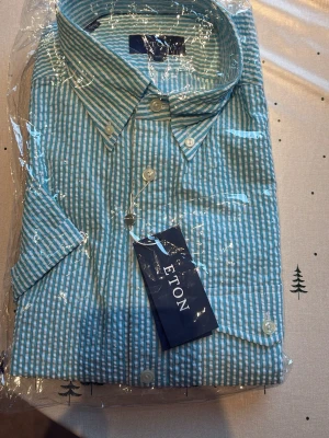 Randig skjorta från Eton i bomull - Snygg skjorta från Eton med blå och vita ränder. Klassisk krage, kort ärm och knappar framtill. Tillverkad i luftig bomull, perfekt för en fräsch och stilren look. Passar dig som gillar tidlös design och hög kvalitet. Skriv för minsta lilla fråga.