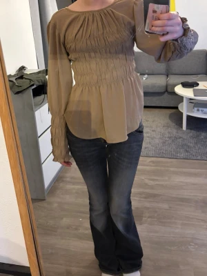 Beige blus med smock och volang - Snygg beige blus med smockdetaljer över bröst och ärmar. Långärmad modell med lätt volang nertill och vid ärmslut. Tunn och luftig i materialet, perfekt för en trendig och avslappnad look.