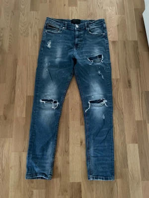 Blå slitna jeans Supply & Demand - Säljer ett par blå jeans från Supply & Demand med slitningar och hål på knäna samt distressed detaljer. Jeansen har fem fickor, knappgylf och en tvättad look med ljusa partier. Perfekta för en avslappnad streetwear-stil.