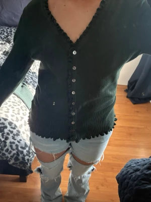 Mörkgrön kofta med volang från Flash Woman - Snygg mörkgrön kofta från Flash Woman i storlek L. Koftan har volangdetaljer längs v-ringningen och nederkanten, samt knappar framtill. Långärmad och tillverkad i mjukt stickat material som känns skönt mot huden. Det står storlek L men den passar mer en M.