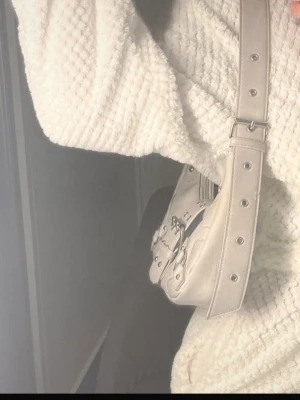 Beige axelväska med spännen - Trendig beige axelväska i en mjuk, rundad form med coola silverfärgade detaljer och dekorativa spännen framtill. Väskan har bred axelrem med nitar och är tillverkad i ett material som liknar konstläder. Perfekt för dig som gillar en stilren men edgy look.