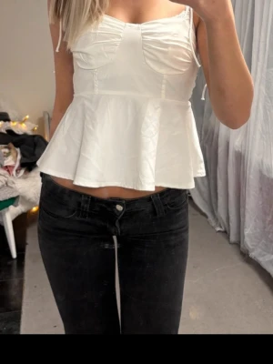 Vit peplum linnetopp med smala band - Snygg vit linnetopp med peplumskärning och smala axelband som knyts. Toppen har markerade sömmar vid bysten och en lätt utsvängd nederdel. Perfekt för dig som vill ha en feminin och trendig look.