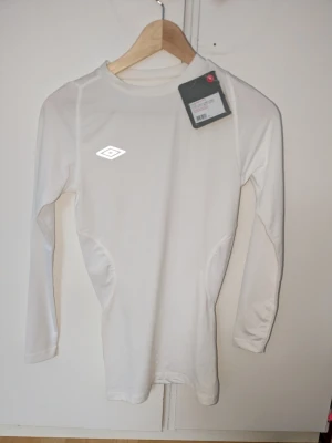 Träningströja - Säljer en vit långärmad träningströja från Umbro. Tröjan har en tight passform och är tillverkad i ett stretchigt funktionsmaterial som passar perfekt för sport och träning. Nypris 399 kr☺️