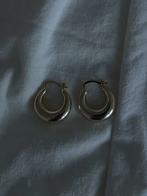 Guldfärgade hoops örhängen - Säljer ett par chunky hoops örhängen i guldfärgat. De har en rund, tjock form och stängs med klassiskt lås. Perfekta för dig som gillar statement-smycken och vill addera lite extra bling till din look.