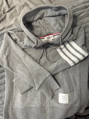 Grå hoodie från Thom Browne - Snygg grå hoodie från Thom Browne med dragkedja, huva med dragsko och vita ränder på ena ärmen. Klassisk patch med logga på magen och fickor framtill. Tillverkad i mjuk bomullsblandning för skön känsla och stilren look. Passar M!