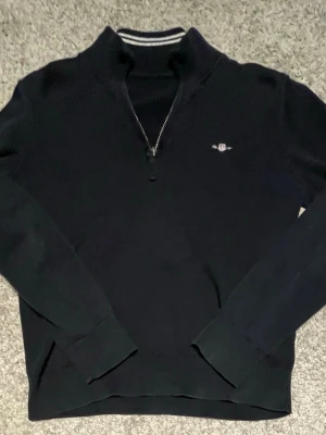 Svart halvzip tröja från Morris - Stilren svart långärmad tröja från Morris med halvzip och diskret logga broderad på bröstet. Tröjan har ribbade muddar vid ärmslut och nederkant samt en klassisk ståkrage med randig detalj på insidan. Perfekt för en snygg och avslappnad look.