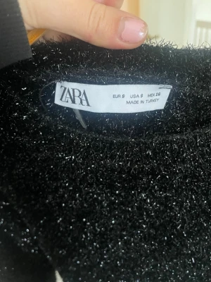 Glittrig svart stickad tröja Zara - Säljer en svart stickad tröja från Zara med glittriga inslag. Tröjan har lång ärm och en klassisk rund halsringning. Materialet är mjukt och fluffigt med en skimrande finish som gör att den sticker ut.