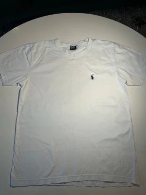 Vit t-shirt från Polo Ralph Lauren - Klassisk vit t-shirt från Polo Ralph Lauren med rund hals och korta ärmar. Tillverkad i 100% bomull och har den ikoniska lilla broderade loggan på bröstet. Perfekt basplagg för en clean och enkel stil.