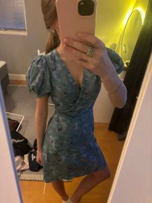 Klänning från Adoore! - Jättefin kort blå klänning med blommigt mönster och puffärmar. Omlottmodell med v-ringning och markerad midja. Perfekt för sommarens alla tillfällen. Nypris ca 1500!