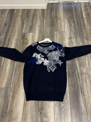 Mörkblå Louis Vuitton tröja - Snygg mörkblå tröja från Louis Vuitton med unikt mönster i silver, vitt och blått över bröstet och axlarna. Tröjan har rund halsringning och långa ärmar. Perfekt för dig som vill sticka ut med exklusiv design.