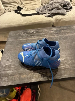 Blåa Puma fotbollsskor med snörning - Säljer ett par blåa fotbollsskor från Puma med vita detaljer och snörning. Skorna har en modern design med röda och vita mönster på sidorna och hälen. Perfekta för dig som vill sticka ut på planen och få bra grepp och kontroll. Gratis för första köpet av något på min sida