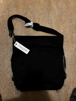 Svart axelväska från Ceannis - Snygg svart axelväska från Ceannis i ett ribbat tygmaterial, helt oanvänd. Väskan har en bred axelrem och enkel, stilren design som passar till många outfits. Perfekt för dig som vill ha en praktisk och trendig väska med plats för det viktigaste. Orginalpris :800kr