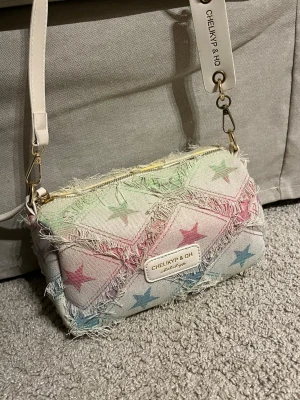 Pastellfärgad stjärnmönstrad handväska - Säljer en unik handväska från CHELIKYP & QH i denimliknande tyg med pastellfärgade stjärnor i rosa, blått och grönt. Väskan har fransiga kanter, avtagbar axelrem i vitt och detaljer i guld. Aldrig använd!