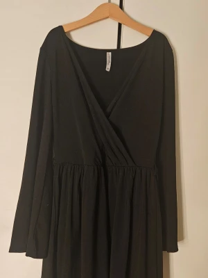 Svart omlottklänning med lång ärm - En svart kortklänning med omlottliv och långa ärmar. Klänningen har en lätt utställd kjol och markerad midja, vilket ger en smickrande siluett. Perfekt för dig som gillar enkel och stilren design.