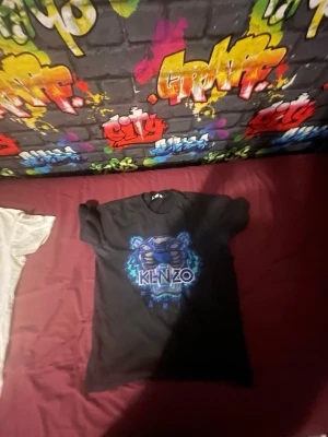 Svart Kenzo t-shirt med tigertryck - Säljer en svart t-shirt från Kenzo med det ikoniska tigertrycket i blått och vitt framtill. T-shirten har rund hals och korta ärmar. Perfekt för dig som vill ha en statement piece i garderoben.