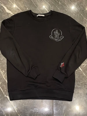 Svart sweatshirt från Moncler - Stilren svart sweatshirt från Moncler med stort broderat logotypmärke på bröstet och en liten detalj i vitt och rött på ärmen. Tröjan har rund halsringning och ribbade muddar vid ärmslut och nederkant. Perfekt för en trendig och avslappnad look. Lite smutsig kring kragen. 