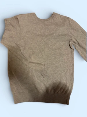 Grå stickad tröja från Hugo Boss - Snygg grå stickad tröja från Hugo Boss med rund halsringning och diskret broderad logga på bröstet. Tröjan har ribbade muddar vid ärmslut och nederkant, perfekt för en stilren och enkel look. Materialet känns mjukt och bekvämt.