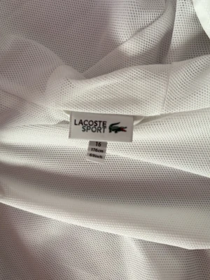 Vit sportjacka från Lacoste Sport - Snygg vit sportjacka från Lacoste Sport i meshmaterial som andas. Jackan har en klassisk design med elastisk nederkant och är perfekt för träning eller uppvärmning. Ikoniska Lacoste-loggan på etiketten. Lätt och bekväm att bära.