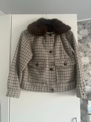 Rutig jacka med fuskpäls-krage - Snygg rutig jacka i beige och brunt med stor, mjuk fuskpäls-krage. Jackan har knappar framtill, två stora fickor och lång ärm. Perfekt för dig som vill ha en trendig och mysig look under höst och vinter.