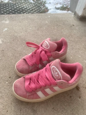 Rosa Adidas sneakers campus  - Säljer ett par snygga rosa Adidas sneakers med klassiska vita ränder på sidorna och breda rosa skosnören. Skorna har låg profil och är tillverkade i mockaliknande material med Adidas-logga på plösen. Perfekta för dig som vill sticka ut med färg på fötterna!
