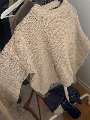 Beige stickad tröja från 157 - Snygg beige stickad tröja från 157 med avslappnad passform och rund halsringning. Tröjan har breda ärmar och ribbstickade muddar vid hals, ärmslut och nederkant. Perfekt för lager-på-lager och enkel att matcha med jeans eller kjol.