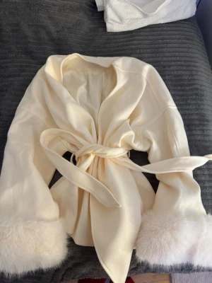 Krämvit kappa med fuskpälsdetaljer - Stilren krämvit kappa med knytskärp i midjan och breda ärmslut dekorerade med fluffig fuskpäls. Kappan har en öppen design utan synliga knappar och är tillverkad i ett mjukt tyg som ger en lyxig känsla. Perfekt för dig som vill ha en elegant och trendig look.