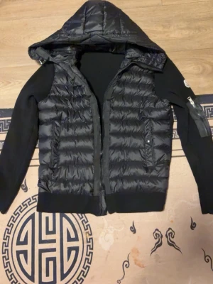 Svart pufferjacka från Moncler - Snygg svart pufferjacka från Moncler med stickade ärmar och huva. Jackan har två framfickor med dragkedja och en extra ficka på ärmen. Moncler-logga på vänster ärm. Perfekt för kalla dagar och riktigt trendig look.
