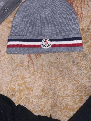 Grå Moncler mössa med ränder - Snygg grå stickad Moncler mössa med klassiska ränder i vitt, rött och marinblått vid kanten. Mössan har Moncler-logga framtill och är tillverkad i mjukt material som håller dig varm. Perfekt accessoar för kyliga dagar.