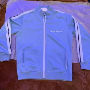 Blå ziptröja från Palm Angels - Snygg blå ziptröja från Palm Angels med vita och svarta ränder längs ärmarna. Tröjan har hög krage, två fickor med dragkedja och vit logga på bröstet. Perfekt för en sportig och trendig look.