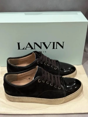 Svarta Lanvin sneakers i mocka och lack - Snygga svarta sneakers från Lanvin med ovandel i mocka och glansig lackad tå. Skorna har bruna skosnören, beige innersula och en vit sula med vintagekänsla. Perfekta för dig som gillar stilrena och exklusiva detaljer.