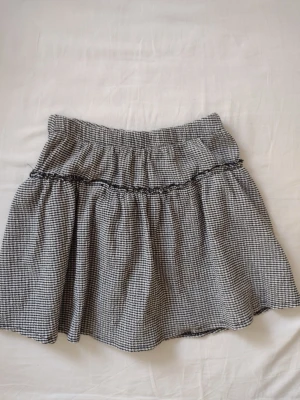 Svartvit rutig kortkjol med volang - Supersöt svartvit rutig kortkjol med volangdetalj och resår i midjan. Kjolen har ett småmönstrat rutigt tyg och är luftig och flowy, perfekt för en chill och trendig look. Passar till både sneakers och boots.