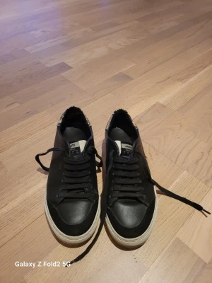 Axel Arigato sneakers  - Snygga svarta sneakers från Axel Arigato med vit sula och detaljer i ormmönstrat material vid hälen. Klassisk låg modell med snörning och diskret logga på sidan. Perfekta för en stilren och trendig look.