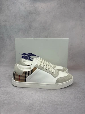 Burberry vita sneakers med rutigt mönster - Snygga vita sneakers från Burberry med klassiskt beige, svart och rött rutigt mönster på sidorna. Skorna har detaljer i ljusgrå mocka, vita skosnören och en vit gummisula. Perfekta för dig som vill ha en stilren och trendig look.