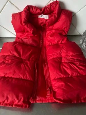 Röd dunväst från H&M i storlek 140 - Snygg röd dunväst från H&M med hög krage och dragkedja framtill. Västen är vadderad och vattenavvisande, perfekt för kyliga dagar. Klassisk quiltad design som ger en sportig look. Märkt med 'Functional Wear' på insidan.