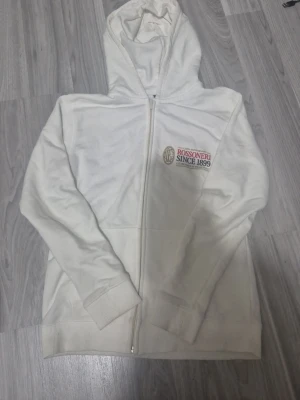 Vit Ac milan zip hoodie - Säljer en vit hoodie med dragkedja från Rossoneri. På bröstet finns ett tryck med texten 'ROSSONERI SINCE 1899' och klubbmärke. Baksidan har stort tryck med text och grafik som hyllar klubbens historia. Hoodien har huva och ribbade muddar. Perfekt för dig som gillar fotboll och stilren design. Tröjan har dock en litwn fläck på ärmen, men det går troligen bort med tvätten