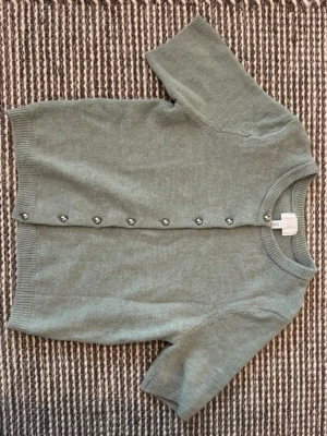 Grön kortärmad kofta från H&M - Kortärmad kofta i mjukt stickat material från H&M. Färgen är dämpat grön och koftan har rund halsringning, ribbade kanter och knappar framtill. Perfekt att slänga över en topp för en chill look.