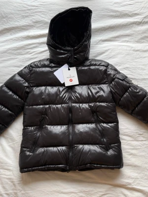 Svart pufferjacka från Moncler/Moncler Maya - Stilren och trendig kort pufferjacka i svart med glansig finish. Jackan har en tidlös design och en rak, modern silhuett som slutar vid höften. Lätt att matcha både till vardag och mer uppklädda outfits.  Jackan är fodrad med dun/fjädrar för bra värme under kyligare dagar och har klassisk quiltning. Utrustad med avtagbar huva, dragkedja framtill samt tryckknappar för extra skydd. Praktiska dragkedjefickor framtill och justerbar nederkant med dragsko för bättre passform. Jackan har en insydd QR-/