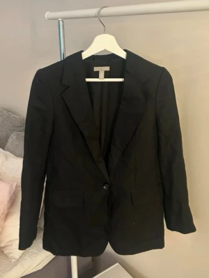 Svart kavaj med klassisk passform - Stilren svart linne kavaj med en knapp framtill och två fickor med lock. Klassisk krage och figurnära passform som ger en snygg siluett. Perfekt att matcha med skjorta eller t-shirt för en clean look. Materialet ser ut att vara bomull eller en bomullsblandning.