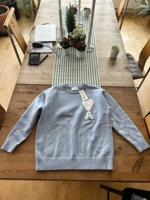 Ljusblå stickad tröja från Ami Paris - Säljer en ljusblå stickad tröja från Ami Paris med deras ikoniska hjärta och bokstaven A broderat på bröstet. Tröjan har rund hals, ribbade muddar och nederkant. Perfekt för dig som gillar stilrena och trendiga plagg.