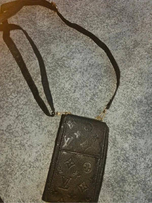 Svart plånbok med axelrem - Snygg svart plånbok i präglat läder med klassiskt monogrammönster. Plånboken har en avtagbar axelrem, flera fack för kort och mynt, samt dragkedja i silverfärg. Perfekt för dig som vill ha både stil och funktion.