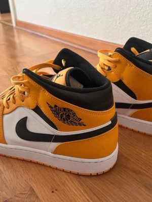 Air Jordan 1 Mid svart/gul/vit - Säljer ett par Air Jordan 1 Mid sneakers i svart, gul och vit. Klassisk siluett med svart swoosh, gul ovandel och vita paneler. Skorna har svart innersula och snörning i gult. Perfekta för dig som vill sticka ut med en ikonisk och sportig look.