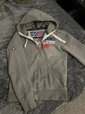 Grå Superdry hoodie med dragkedja - Säljer en grå hoodie från Superdry med dragkedja, huva och vita dragsnören. Broderad Superdry-logga i blått, vitt och rött på bröstet samt en patch på ärmen. Mjuk insida och kängurufickor framtill. Perfekt för en sportig och avslappnad stil.
