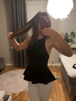 Svart peplum topp med halterneck - Snygg svart peplum topp med halterneck och ärmlös design. Toppen har en figurnära passform upptill och volang nedtill som ger en feminin siluett. Perfekt att styla med både jeans och kjol för en trendig look.