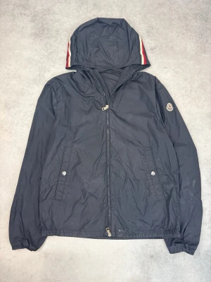 Svart vindjacka från Moncler - Snygg svart vindjacka från Moncler med huva och dragkedja framtill. Jackan har Moncler-logga på ärmen och röda/vita ränder på huvan. Två fickor med knappstängning och elastiska muddar. Tillverkad i lätt, vindtätt material.