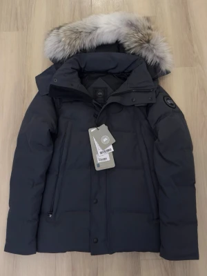 Mörkgrå jacka från Canada Goose - Säljer en svart dunjacka från Canada Goose med klassisk patch på ärmen. Jackan har huva, dragkedja och tryckknappar framtill. Tjockt vadderad för extra värme och perfekt för kalla vinterdagar. Snygg och tidlös design som funkar till det mesta. ( WYNDHAM PARKA) kvitto finns med 