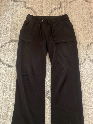 Svarta mjukisbyxor med raka ben - Säljer ett par svarta mjukisbyxor med raka ben och resår i midjan med dragsko. Byxorna har två stora fickor framtill och är gjorda i ett mjukt, stretchigt material som känns riktigt skönt. Perfekta för chill dagar eller när du vill vara bekväm.