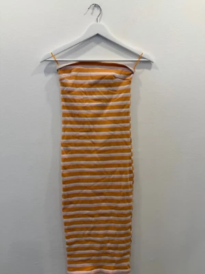 Randig långklänning från Gina Tricot - Säljer en lång, ärmlös klänning från Gina Tricot med smala axelband. Klänningen har ett randigt mönster i orange, rosa och vitt. Materialet är mjukt och stretchigt, perfekt för varma dagar.