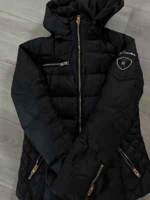 Svart pufferjacka med huva från Rockandblue - Snygg svart pufferjacka från Rockandblue med guldfärgade dragkedjor och flera fickor. Jackan har en stor huva, quiltade detaljer på ärmar och sidor samt Rockandblue-logga på ena ärmen. Perfekt för kalla vinterdagar och riktigt stilren look.